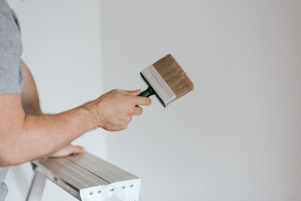 Side view of crop faceless painter in casual clothes holding paintbrush while leaning on stepladder on gray background in